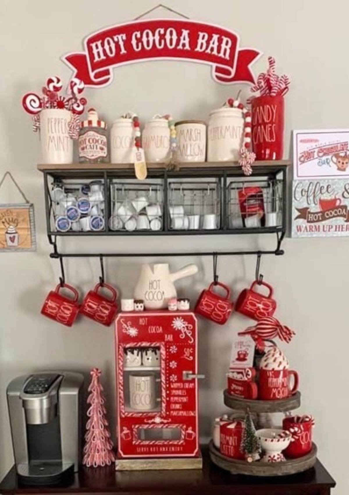 A festive hot cocoa bar with a red and white theme. The bar includes a variety of toppings like marshmallows, candy canes, and chocolate chips. There are also mugs, a coffee machine, and a decorative hot chocolate vending machine.