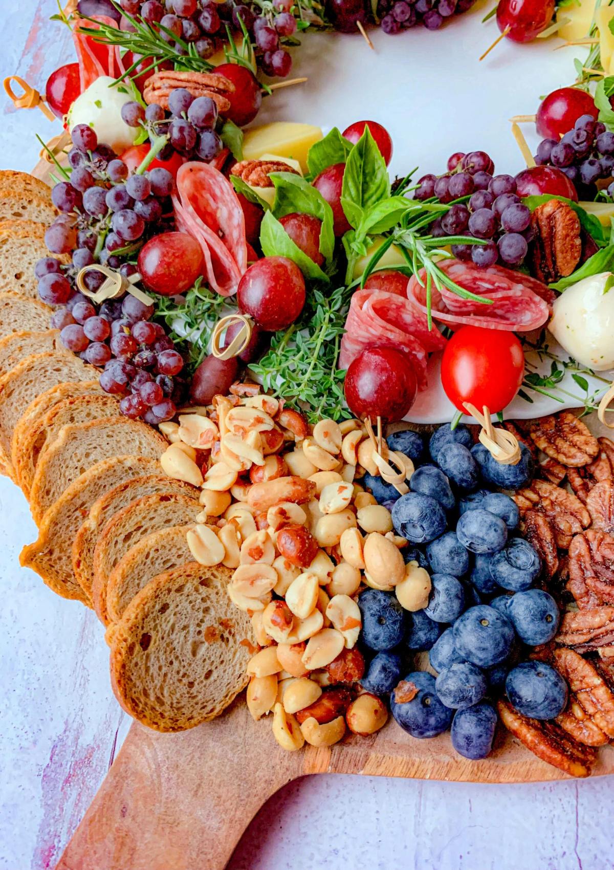 A festive charcuterie board featuring a variety of cheeses, meats, fruits, nuts, and bread. The board is decorated with fresh herbs and grapes.