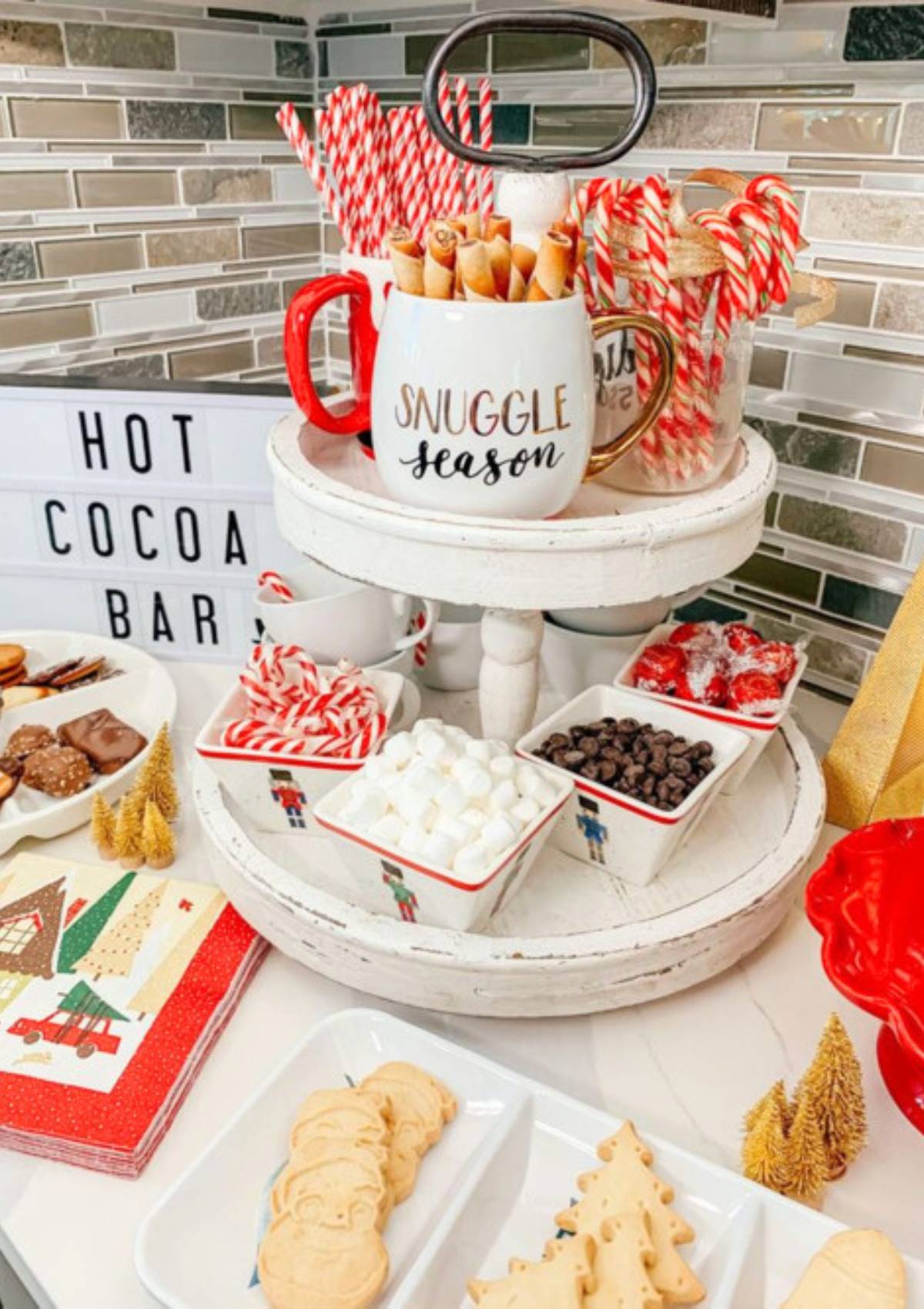 A festive hot cocoa bar with a tiered tray holding various toppings like marshmallows, chocolate chips, and candy canes. There are also mugs, cookies, and a sign that says "Hot Cocoa Bar".