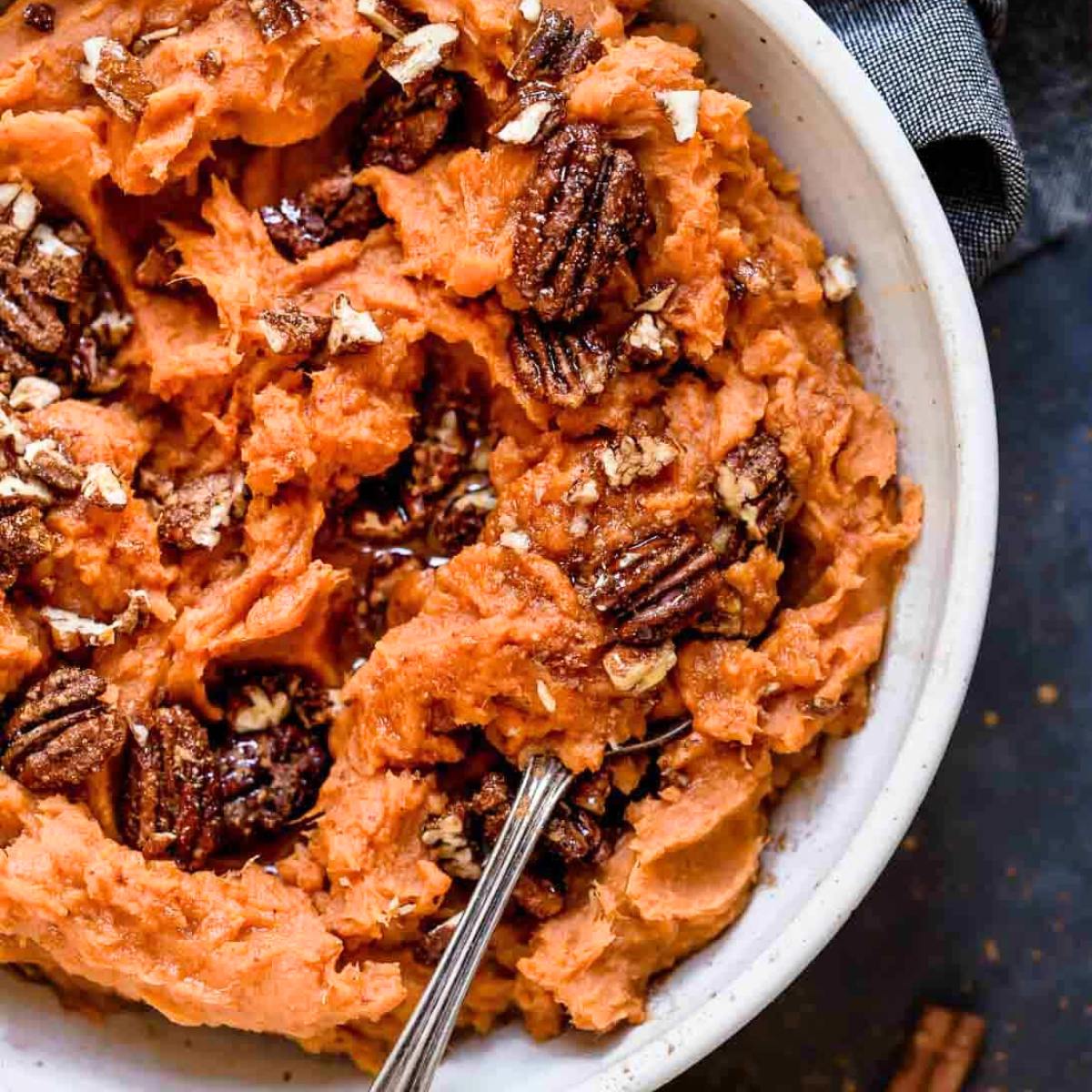 A bowl of creamy mashed sweet potatoes topped with chopped pecans and a drizzle of maple syrup.