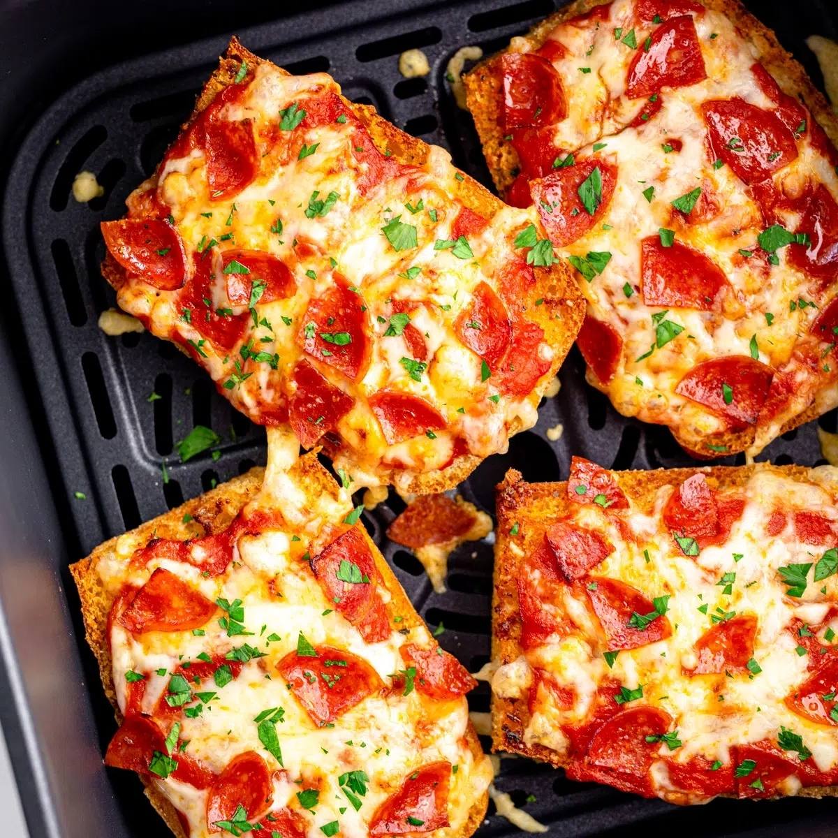 A close-up of mini pizzas cooking in an air fryer basket. The pizzas have melted cheese, pepperoni, and a sprinkle of parsley.