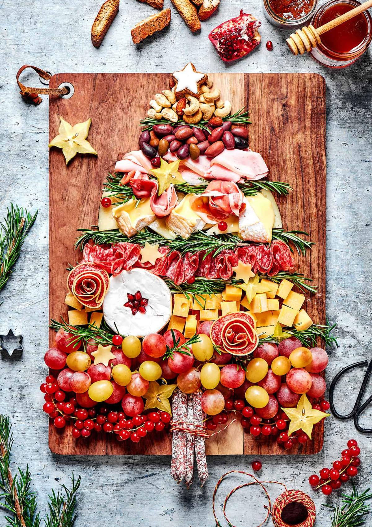 A festive Christmas tree-shaped charcuterie board filled with a variety of cheeses, meats, fruits, and nuts. The board is decorated with rosemary sprigs and star-shaped cookie cutters.