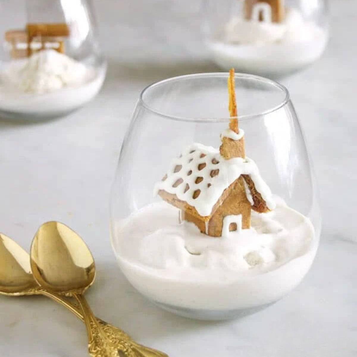 A miniature gingerbread house nestled in a glass filled with a creamy dessert, resembling a snowy scene.