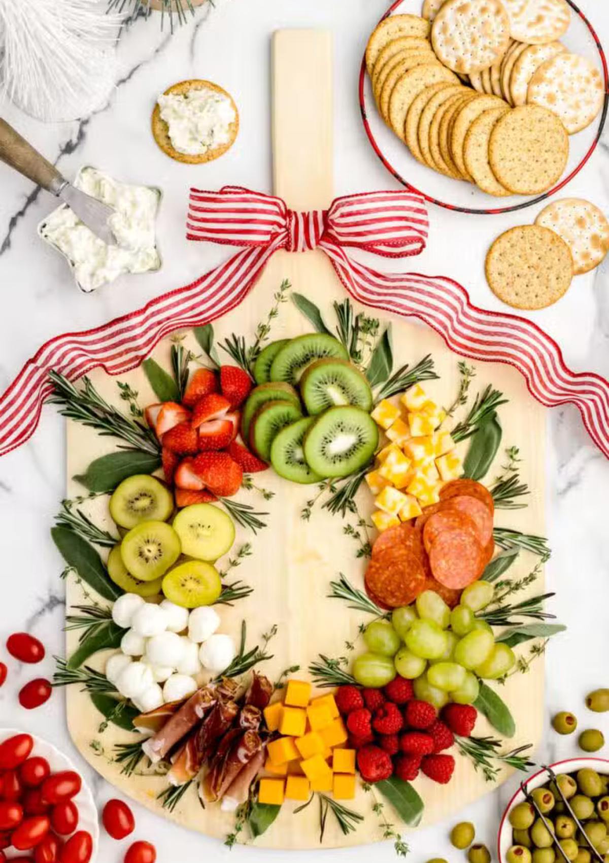 A festive Christmas wreath-shaped charcuterie board filled with a variety of fruits, cheeses, meats, and crackers. The board is decorated with rosemary sprigs and a red ribbon.