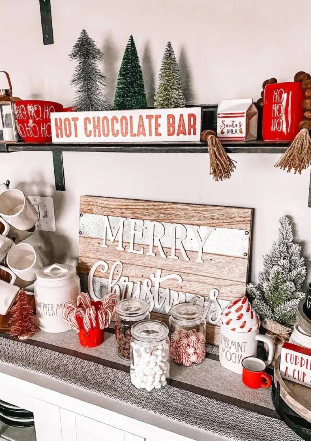 A festive hot cocoa bar with a sign that says "Merry Christmas." The bar includes jars of marshmallows, chocolate chips, and candy canes. There are also mugs, candy canes, and small Christmas trees on display.