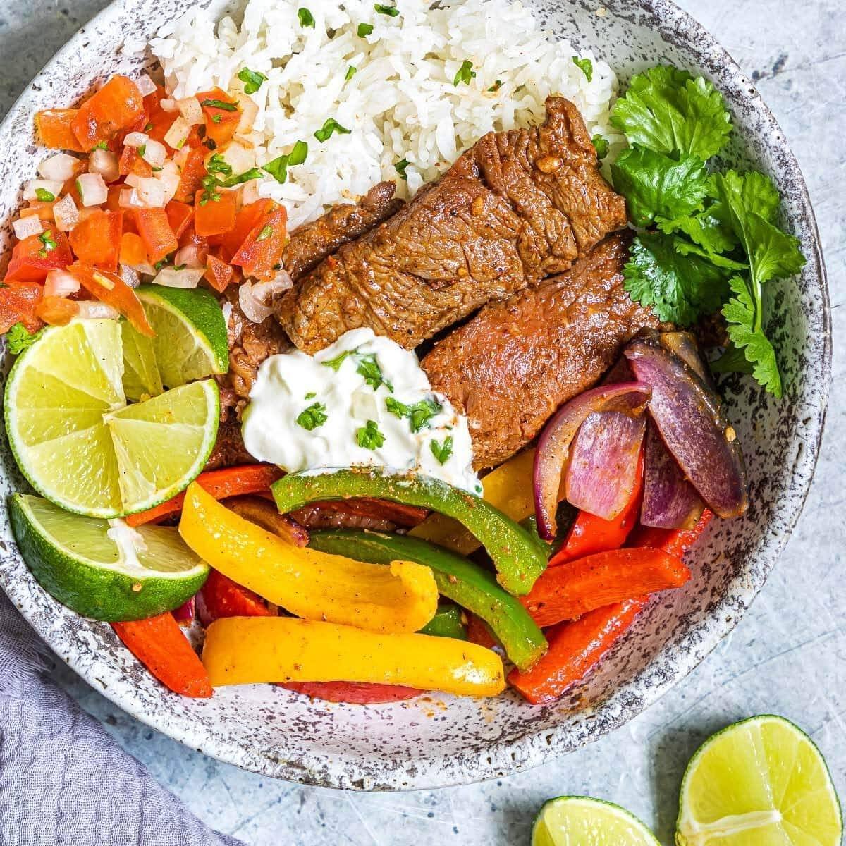 A vibrant and colorful steak fajita bowl. The bowl is filled with seasoned steak, sautéed bell peppers and onions, white rice, pico de gallo, sour cream, and lime wedges.