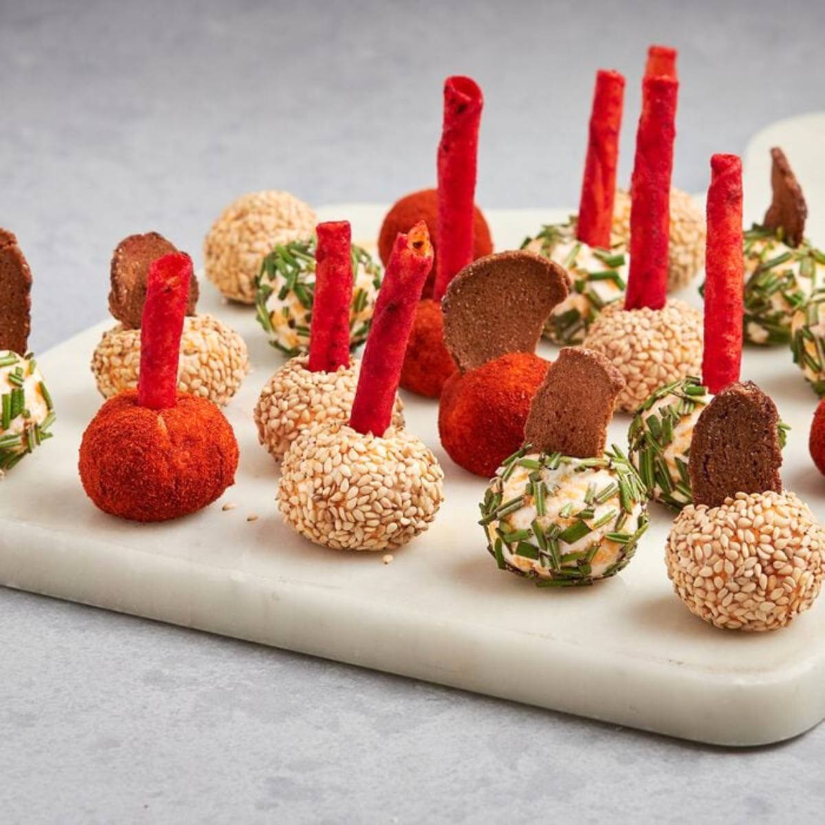 A platter of colorful cheese balls coated in various toppings like sesame seeds, paprika, and herbs. Each cheese ball is topped with a crispy cracker or breadstick.