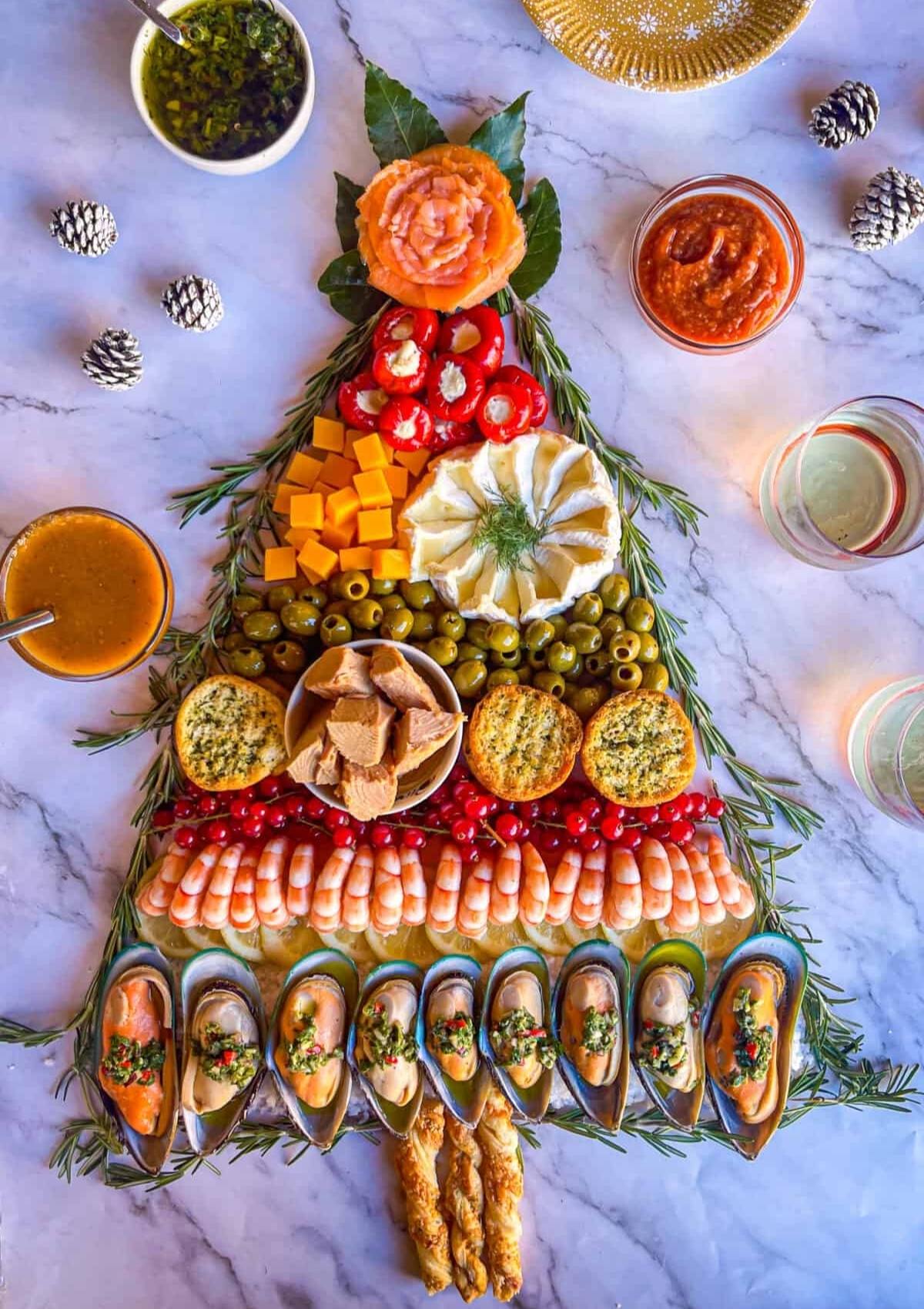 A festive Christmas tree-shaped charcuterie board filled with a variety of seafood, cheeses, olives, and crackers. The board is decorated with rosemary sprigs and small pine cones.