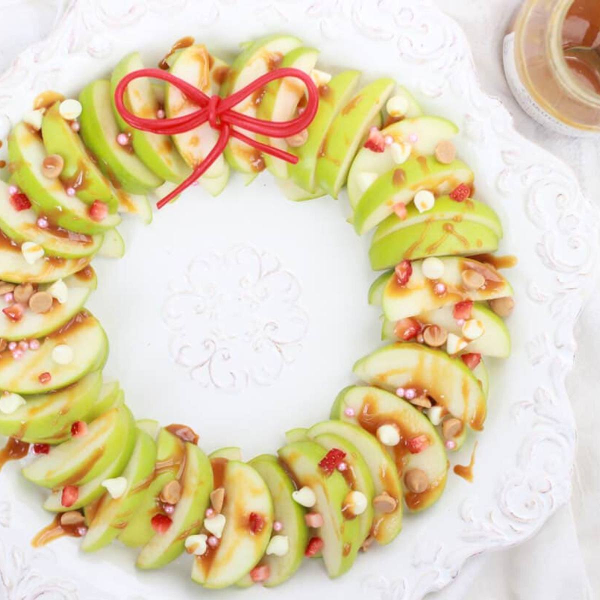 A festive apple wreath arranged on a white plate. The apple slices are drizzled with caramel sauce and topped with chocolate chips, sprinkles, and a red ribbon bow.