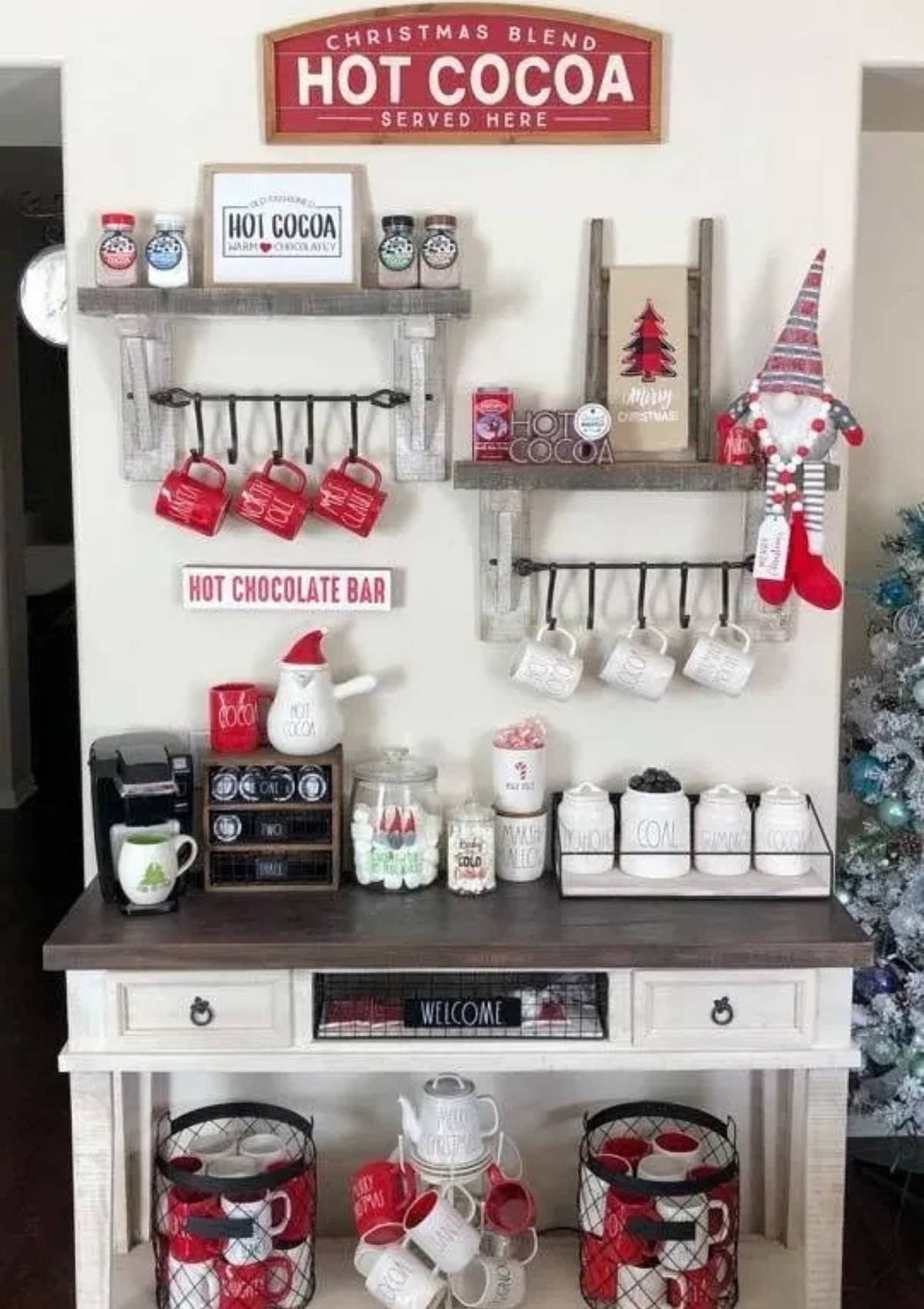 A festive hot cocoa bar with a sign that says "Christmas Blend Hot Cocoa Served Here." The bar includes a variety of mugs, candy canes, marshmallows, and other toppings. There is also a coffee maker and a Christmas tree.