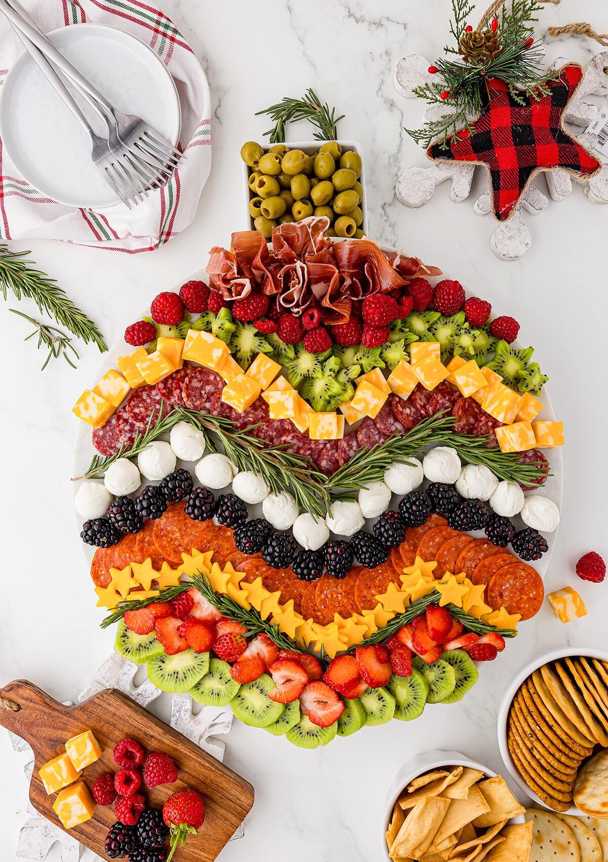 A festive Christmas ornament-shaped charcuterie board filled with a variety of cheeses, meats, fruits, and olives. The board is decorated with rosemary sprigs and a small Christmas ornament.