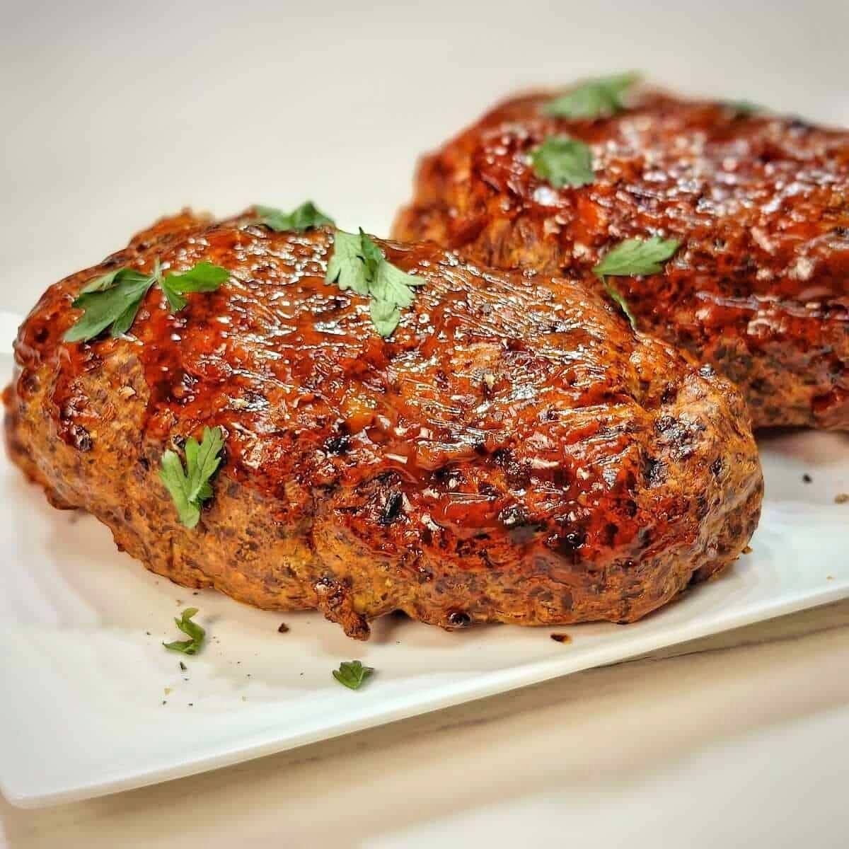 Two golden-brown meatloaves on a white plate. The meatloaves are glazed with a sweet sauce and garnished with fresh parsley.