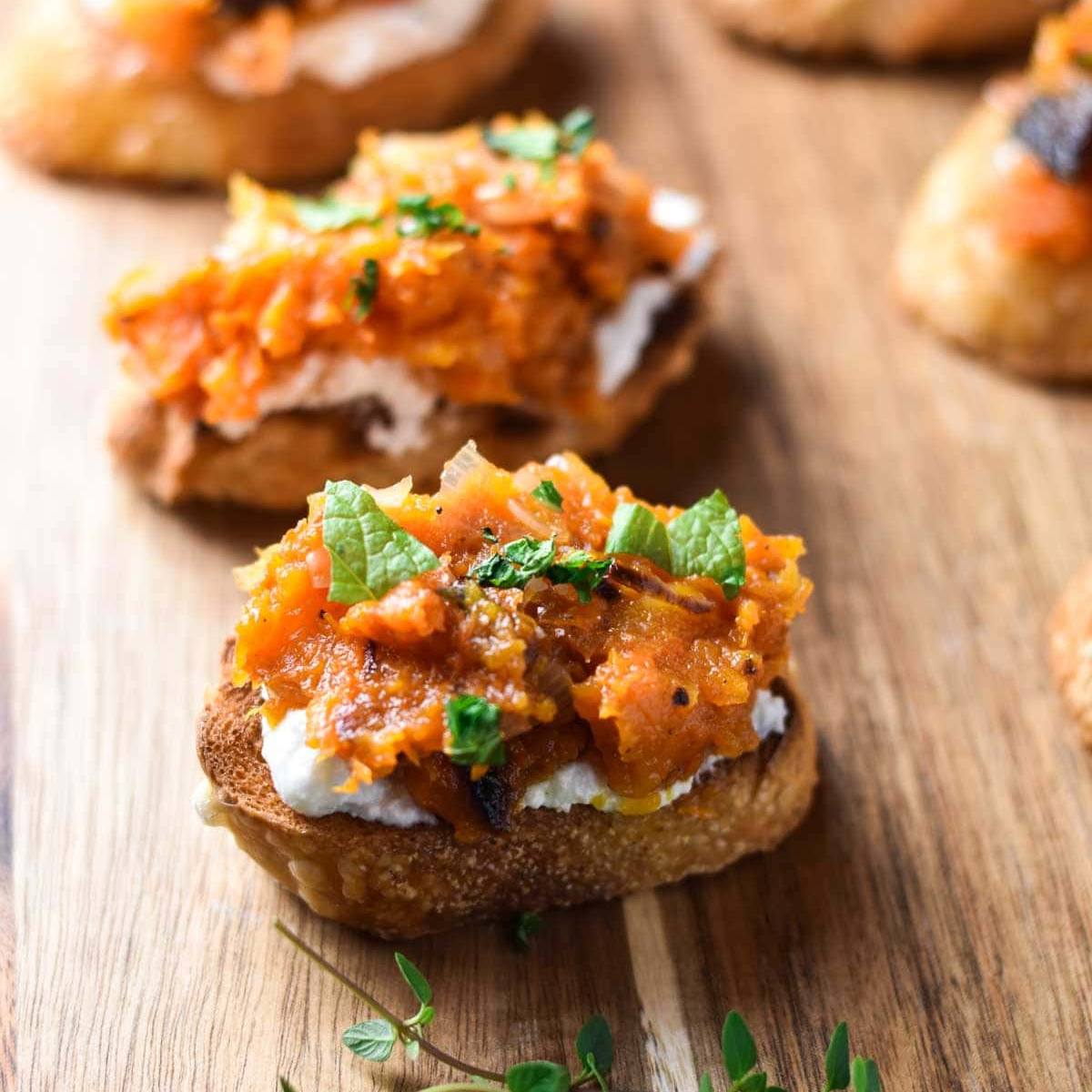 A close-up of crostini topped with roasted butternut squash, ricotta cheese, and fresh thyme leaves.