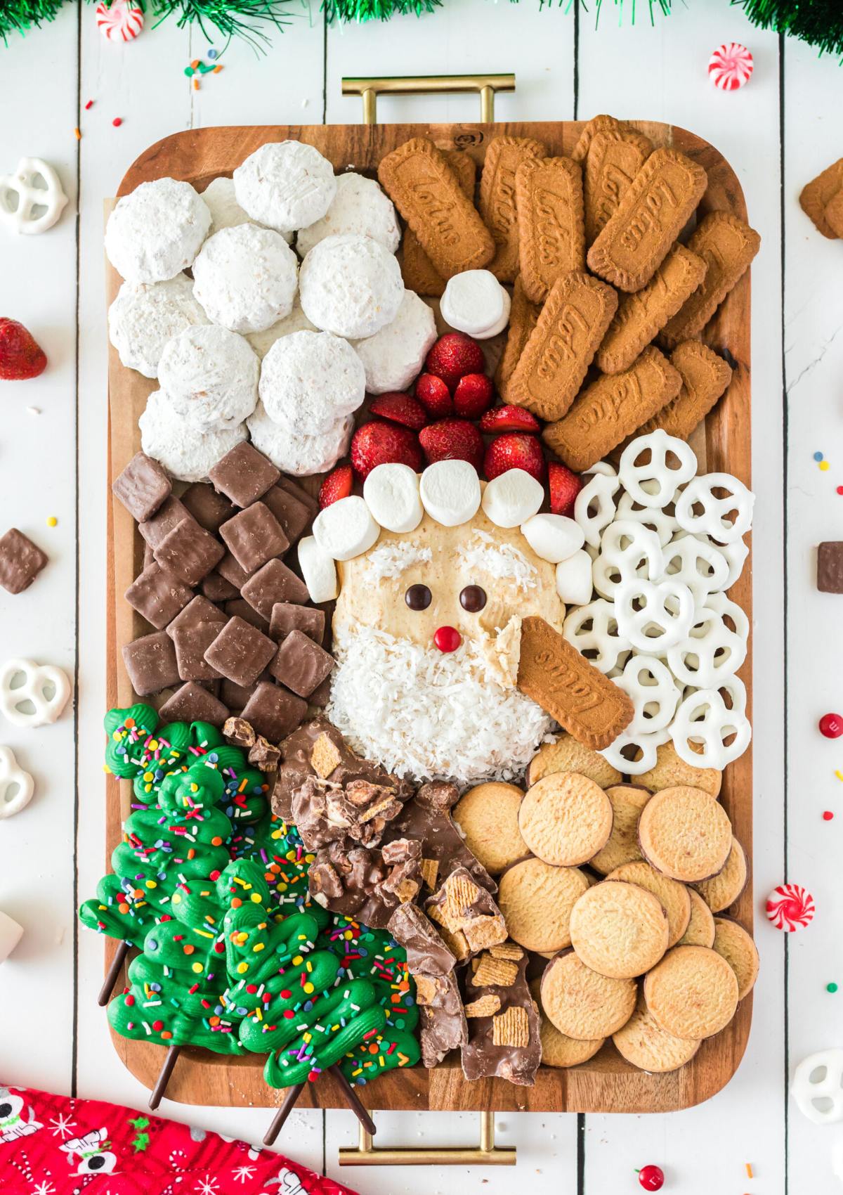 A festive Christmas charcuterie board featuring a Santa Claus made of peanut butter, marshmallows, and pretzels. The board is filled with a variety of other snacks, including cookies, candy, and fruit.