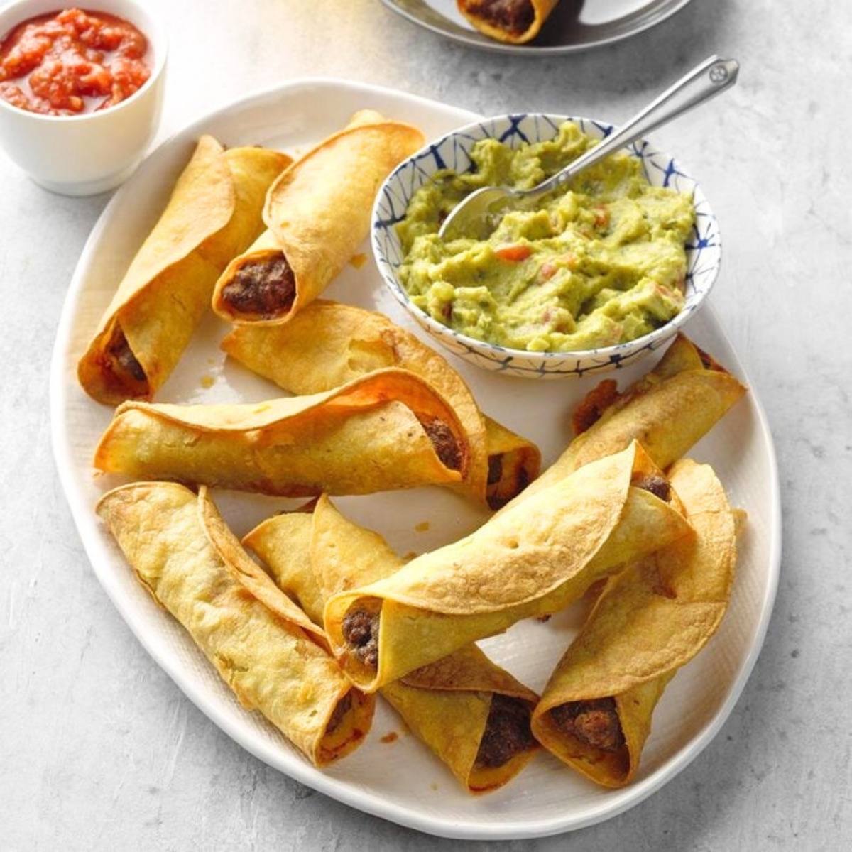 A platter of crispy, golden-brown taquitos arranged neatly. Alongside are bowls of guacamole and salsa, ready for dipping.