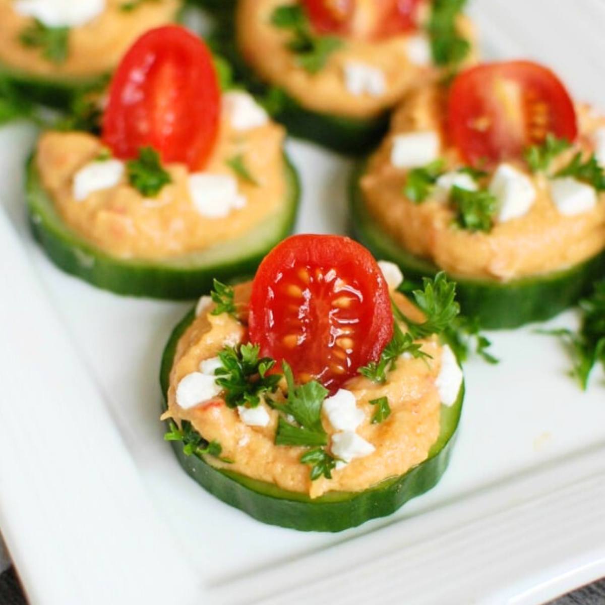 A plate of cucumber slices topped with hummus, feta cheese, cherry tomatoes, and fresh parsley.