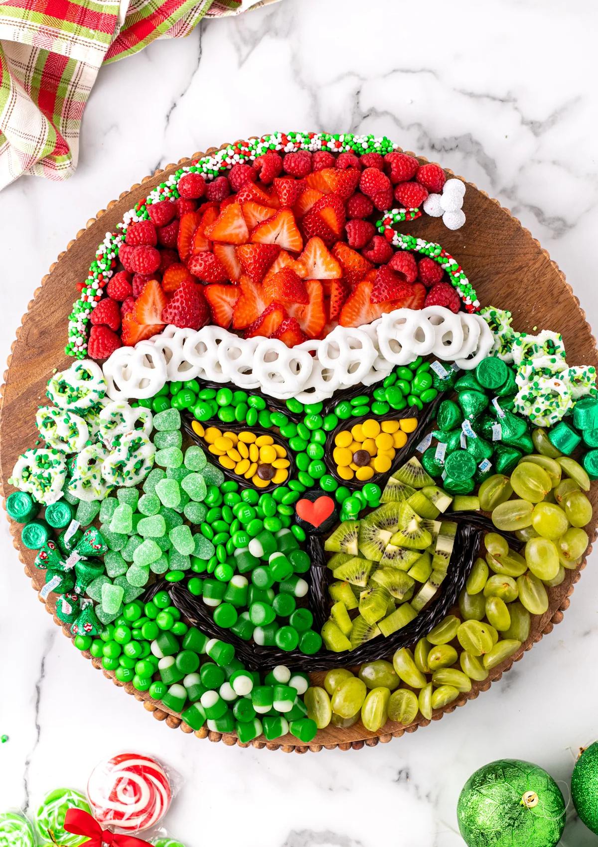 A festive Grinch-themed charcuterie board filled with an assortment of candies, fruits, and snacks. The board features a Grinch face made of green candies, with strawberries and raspberries representing his hat.