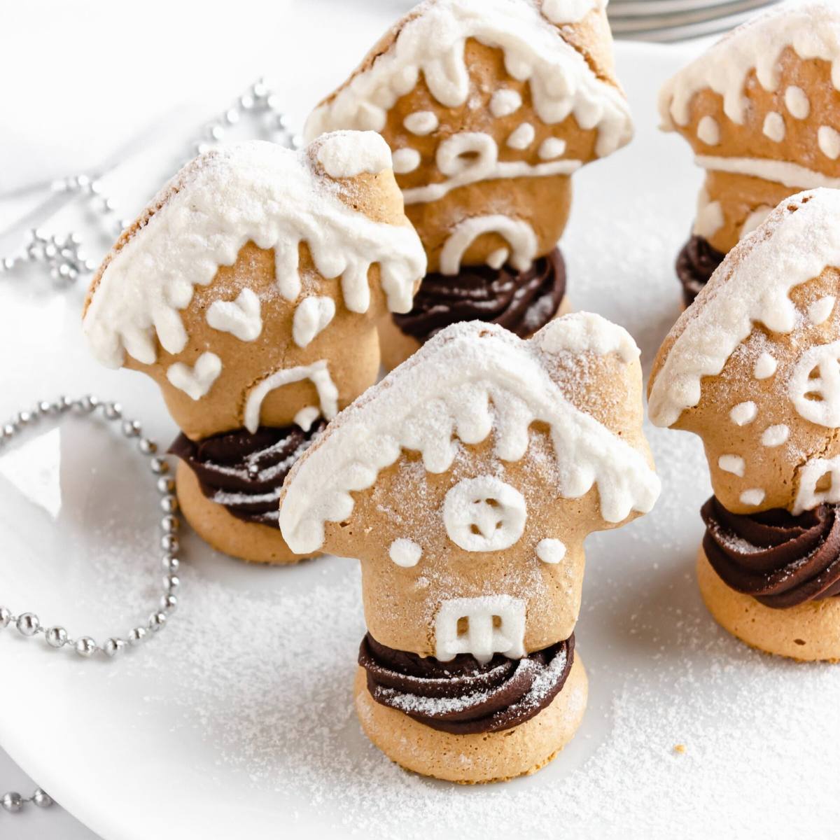 A plate of adorable gingerbread house-shaped macarons. The macarons are decorated with white icing to look like gingerbread houses, complete with doors, windows, and roofs. Some of the macarons are filled with chocolate ganache.