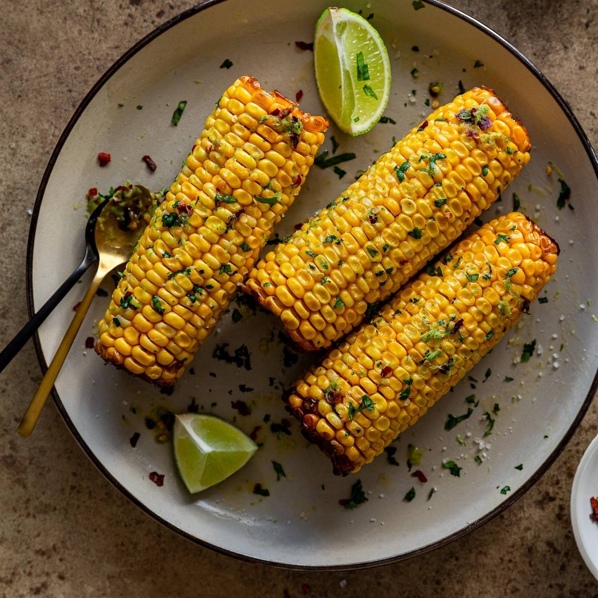 A plate of three air fried corn on the cob. The corn is golden yellow and sprinkled with herbs and spices. There are also lime wedges and a small bowl of a green sauce, possibly a cilantro-lime sauce, on the plate.







