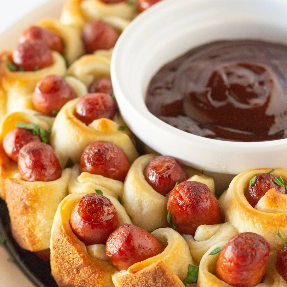 A ring of golden-brown crescent rolls wrapped around small sausages, served with a bowl of barbecue sauce.