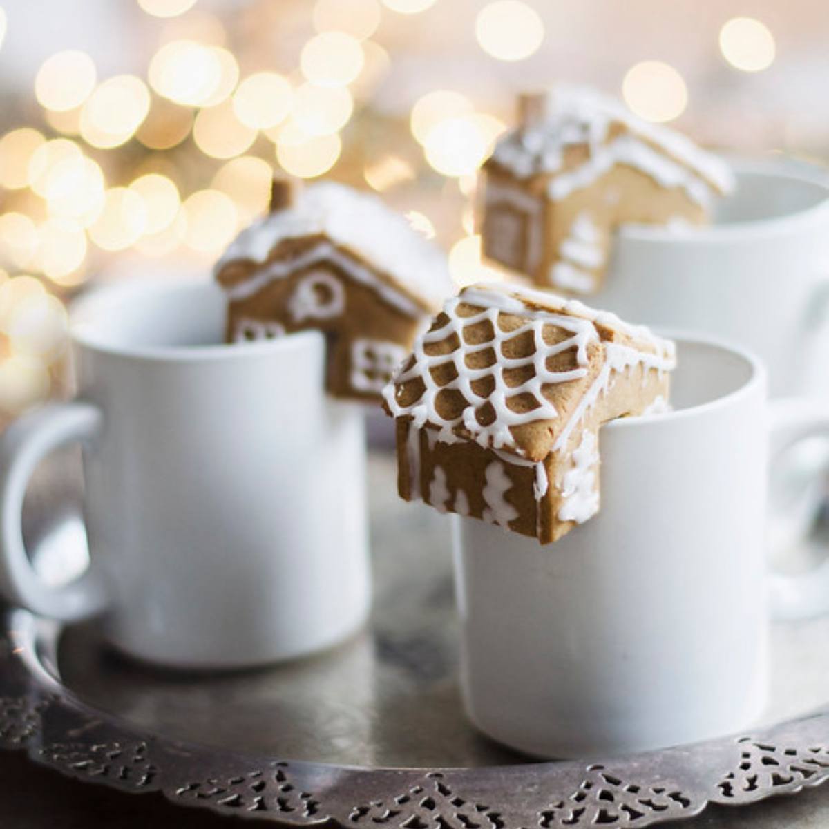 Small gingerbread houses perched on the rims of white mugs, with a festive bokeh background.