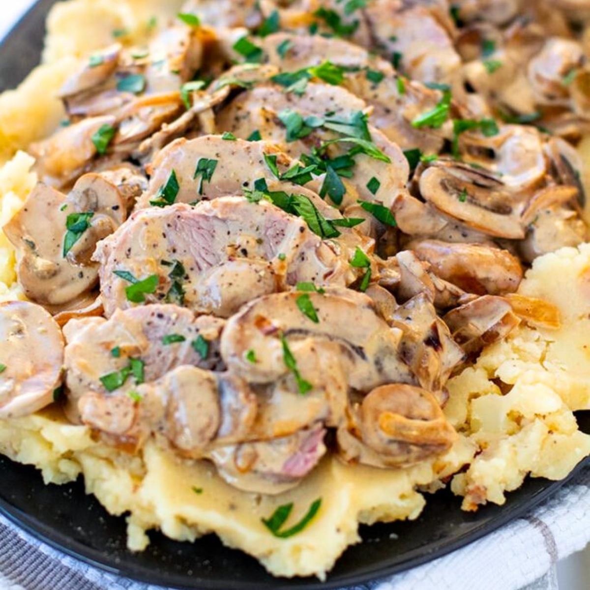 Sliced pork tenderloin covered in a creamy mushroom sauce and served over a bed of mashed potatoes. The dish is garnished with fresh parsley.