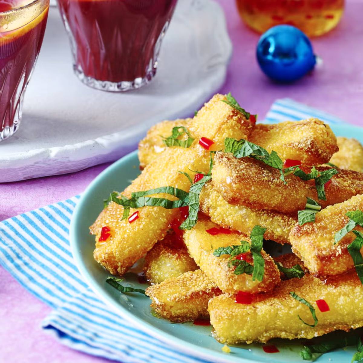 A plate of crispy, golden-brown cornmeal-coated fish fingers, drizzled with a sweet chili sauce and garnished with fresh mint leaves.