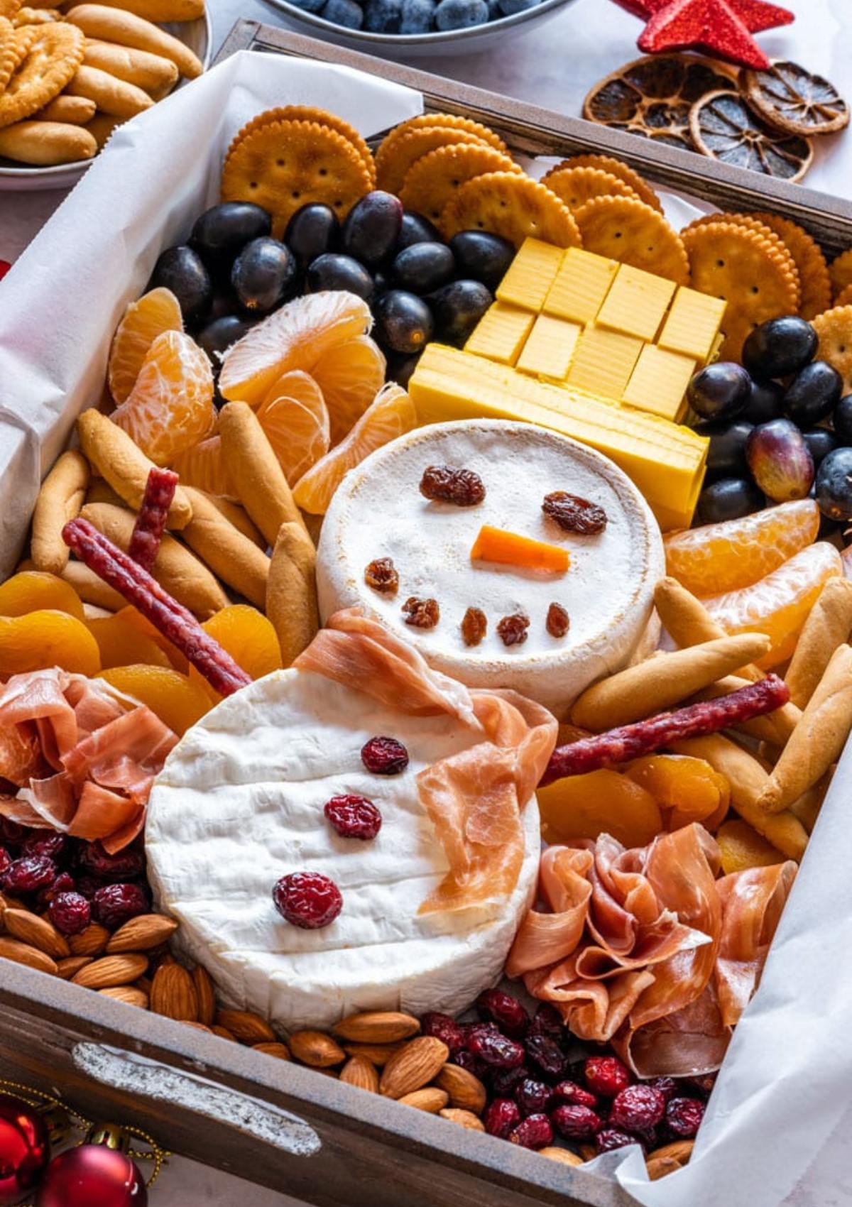 A festive Christmas cheese board featuring a snowman made of cheese, crackers, and dried fruit. The board is filled with a variety of other snacks, including grapes, tangerines, almonds, dried apricots, salami, and breadsticks.