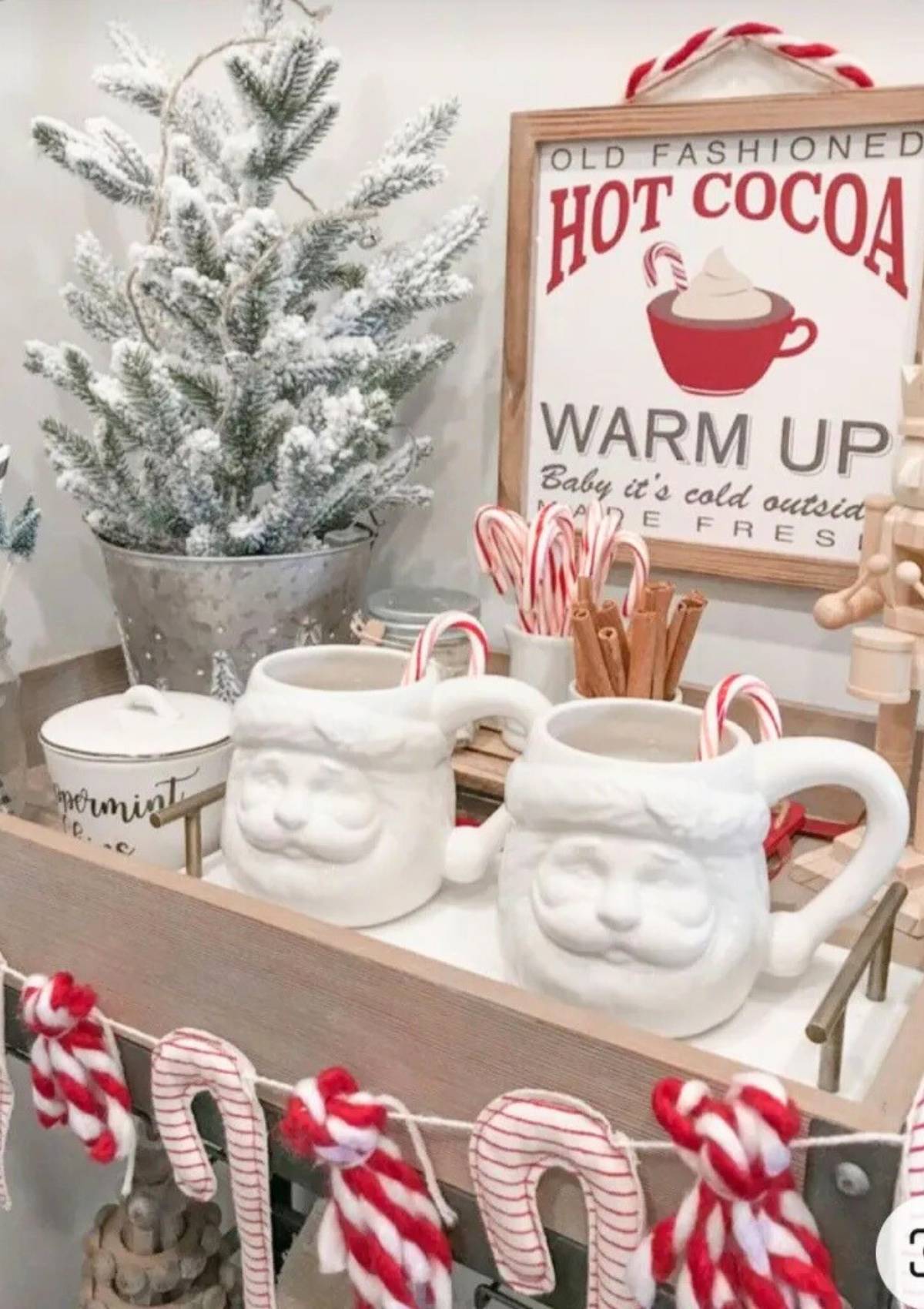 A festive hot cocoa bar with mugs shaped like Santa Claus faces. There are also candy canes, cinnamon sticks, and a framed sign that says "Old Fashioned Hot Cocoa".