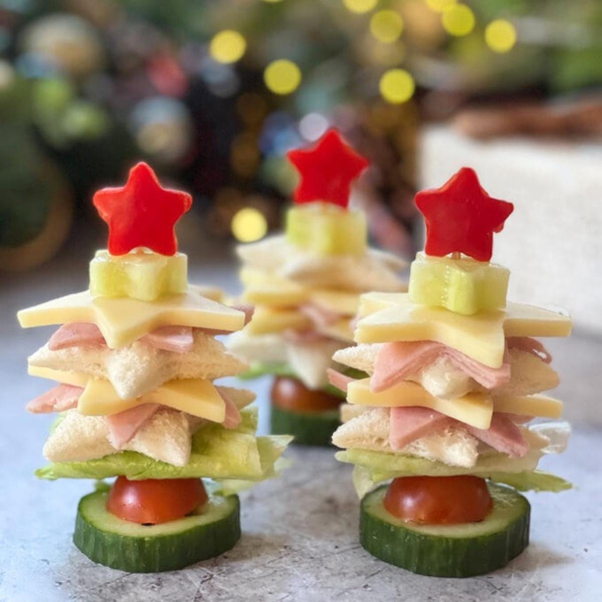 Three festive Christmas tree sandwiches made with layers of bread, ham, cheese, cucumber, and tomato. Each tree is topped with a red star-shaped pepper.
