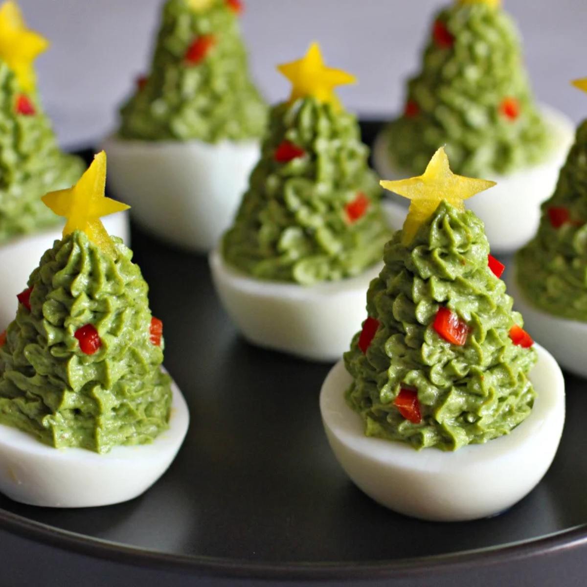 A plate of festive Christmas tree deviled eggs. The eggs are filled with a green avocado filling and decorated with red pepper flakes and yellow pepper stars.