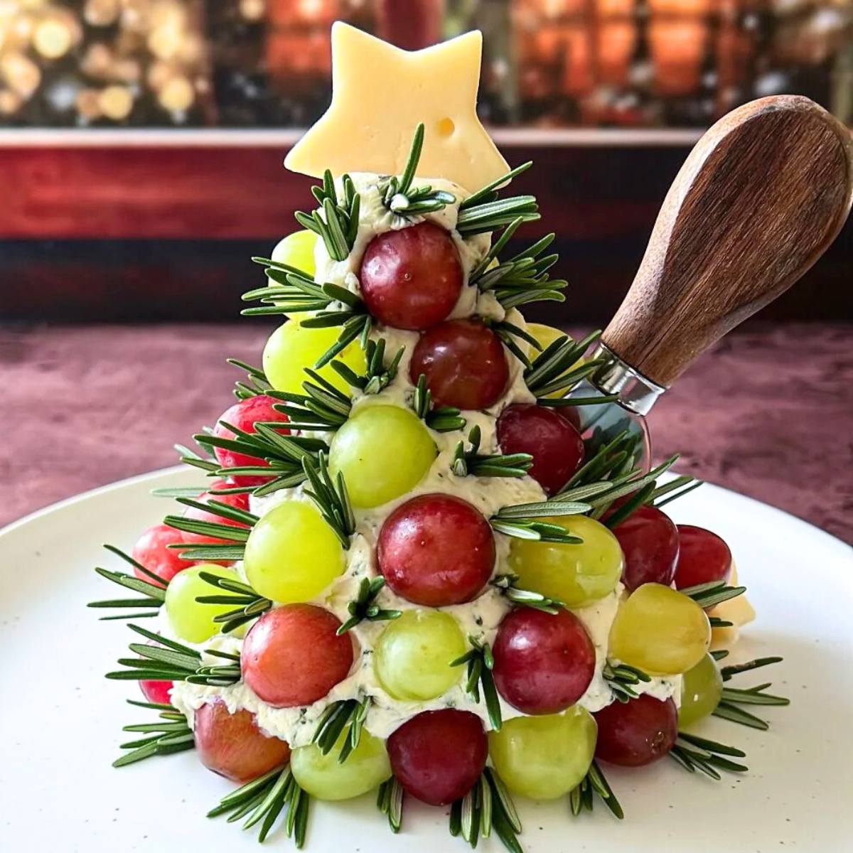 A festive Christmas tree-shaped cheese ball made with cream cheese, herbs, and grapes. The cheese ball is decorated with rosemary sprigs to resemble pine needles and a star-shaped cheese slice on top. A cheese knife is placed next to the tree.