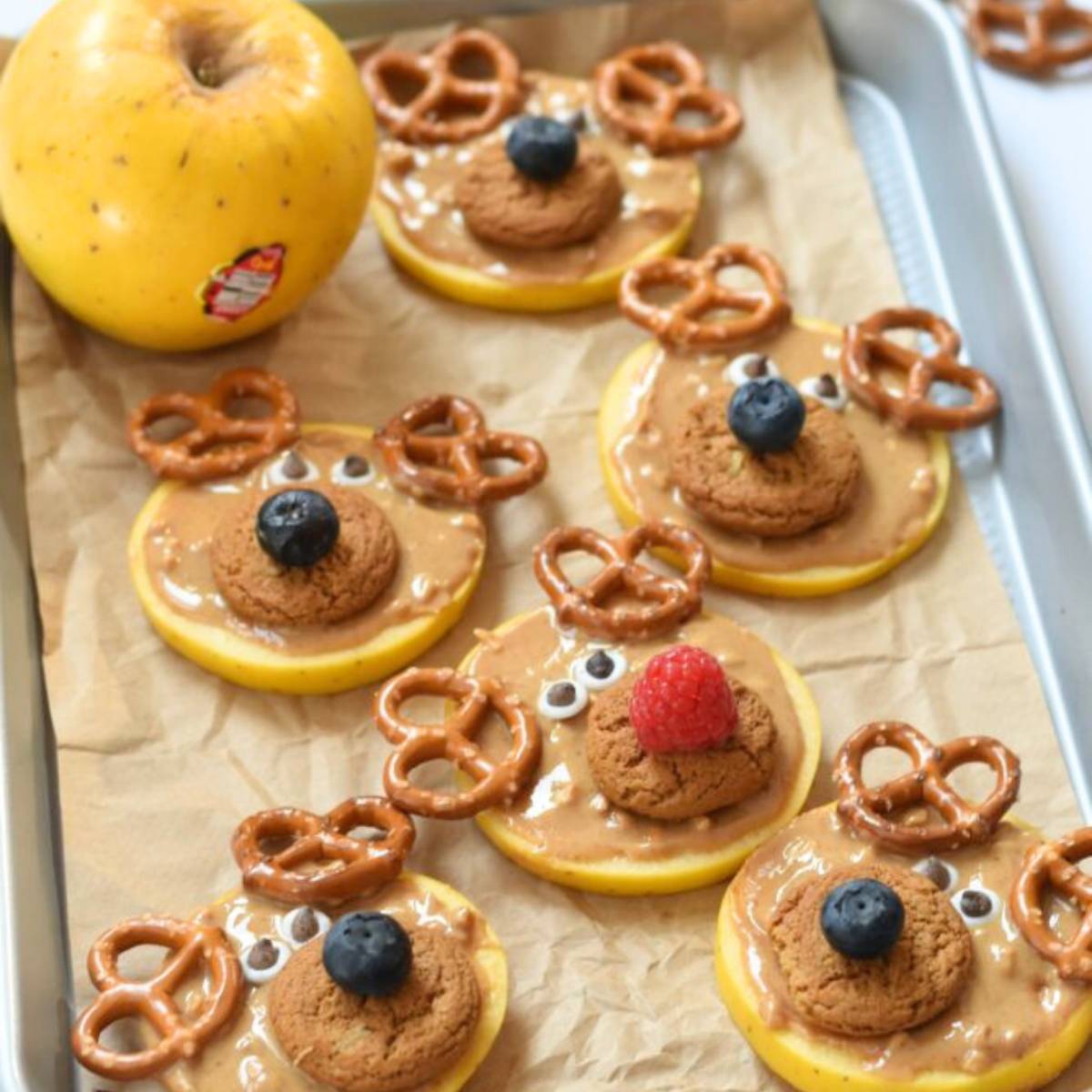 A baking sheet filled with adorable reindeer apple slices. The apple slices are spread with peanut butter and decorated with pretzels as antlers, cookie pieces as noses, and blueberries or raspberries as eyes.