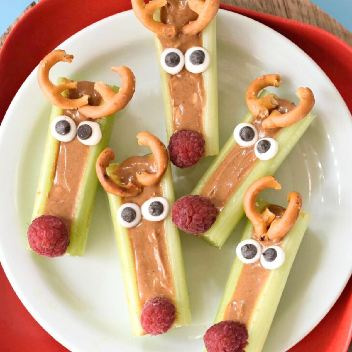 A plate of festive reindeer snacks made with celery sticks. Each celery stick is filled with peanut butter and decorated with pretzels as antlers, chocolate chips as eyes, and raspberries as noses.