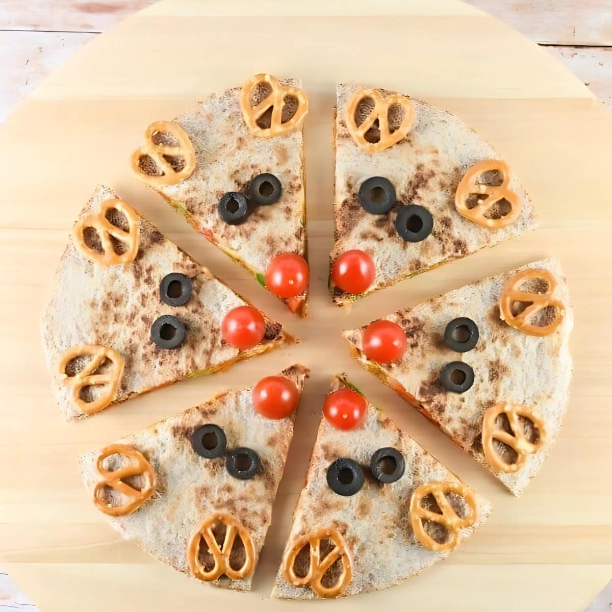 A festive reindeer pizza! The pizza is cut into slices, and each slice has a reindeer face made with olives for eyes, a cherry tomato for a nose, and pretzel sticks for antlers.