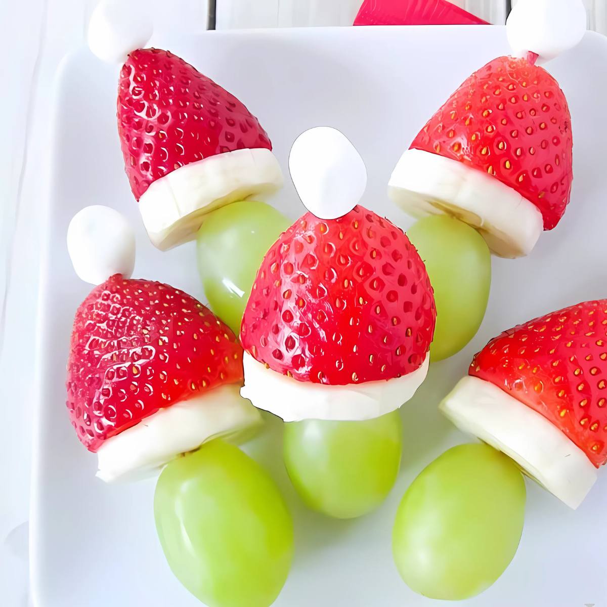 A plate of festive fruit skewers. Each skewer features a strawberry, a grape, and a marshmallow. The strawberry and marshmallow are arranged to resemble Santa hats.