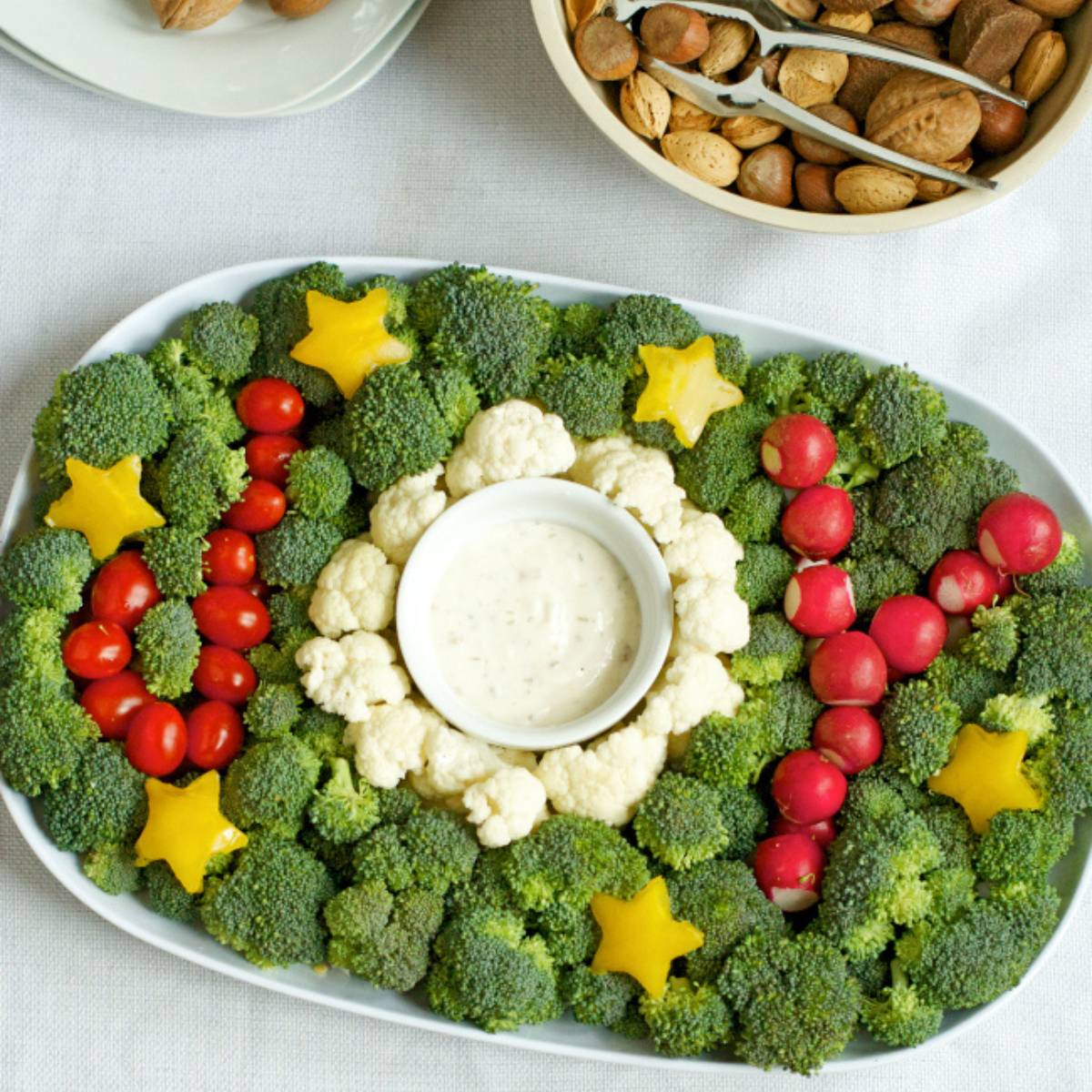 A festive vegetable platter arranged in the shape of a Christmas tree. The platter features broccoli florets, cauliflower florets, cherry tomatoes, and radish roses. A small bowl of dip is placed in the center of the tree.