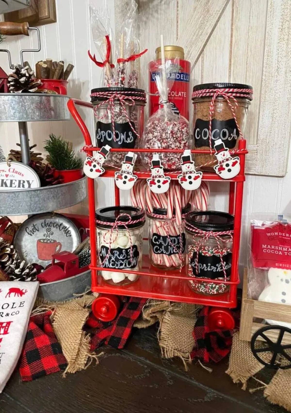 A festive red cart filled with jars of hot chocolate mix, marshmallows, candy canes, and sprinkles.