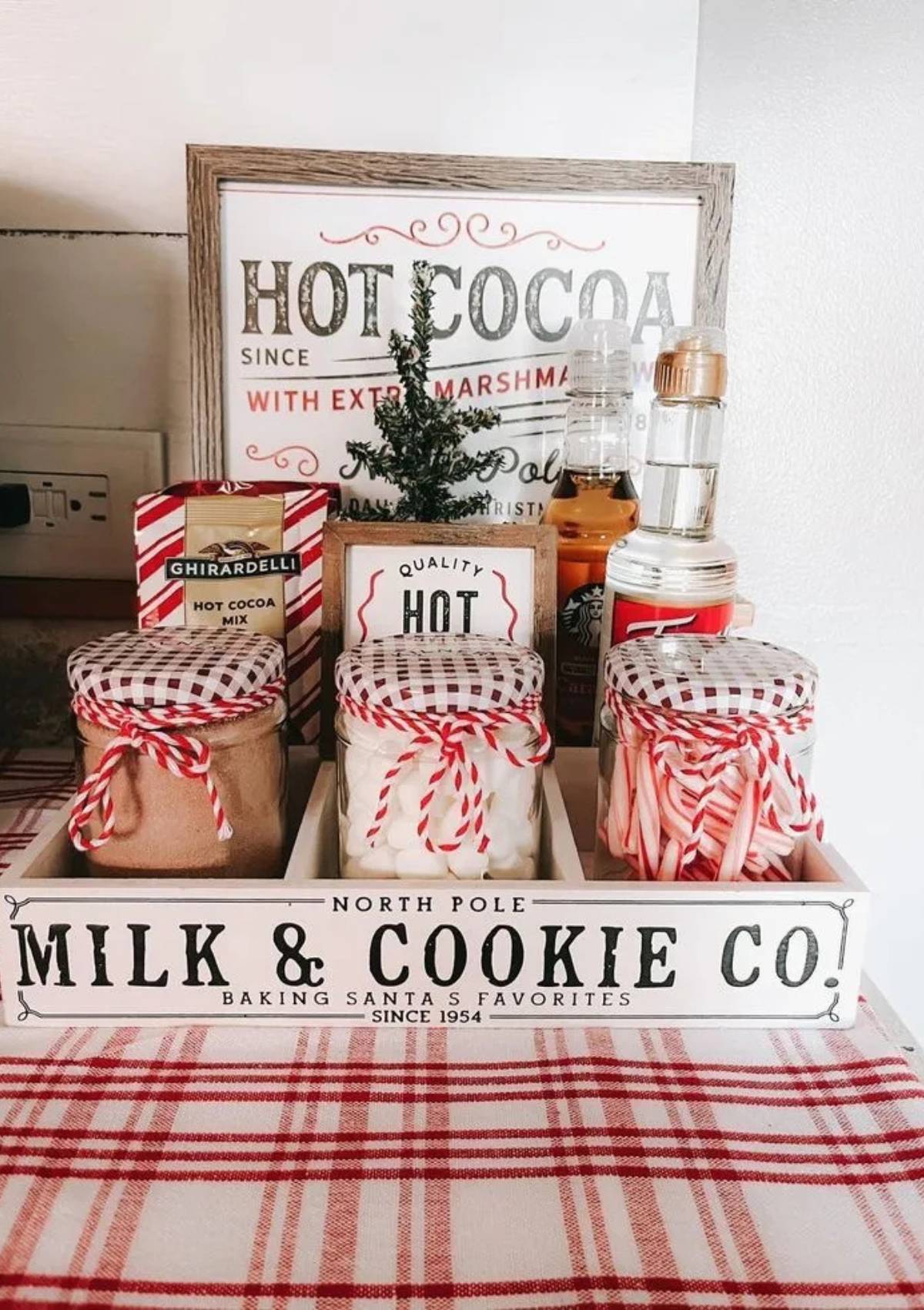 A festive hot cocoa bar set up on a wooden tray. " The bar includes jars of hot cocoa mix, marshmallows, candy canes, and other ingredients for making hot cocoa. There is also a framed sign that says "Hot Cocoa with Extra Marshmallows."