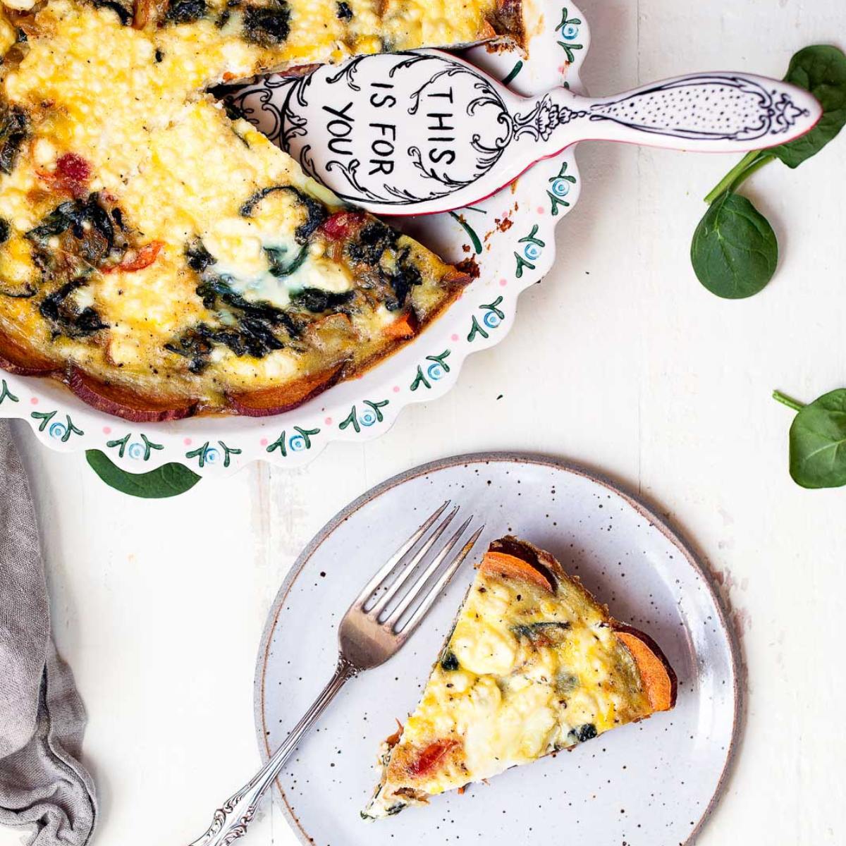 A golden-brown quiche with a sweet potato crust, filled with spinach, tomatoes, and goat cheese. A slice is on a plate, ready to be enjoyed.