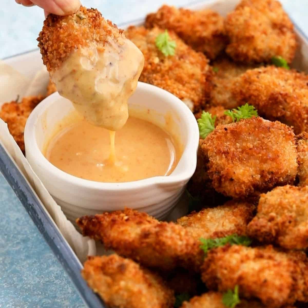 A hand dipping a crispy, golden-brown chicken nugget into a bowl of creamy dipping sauce. The nuggets look delicious and perfect for a snack or meal.