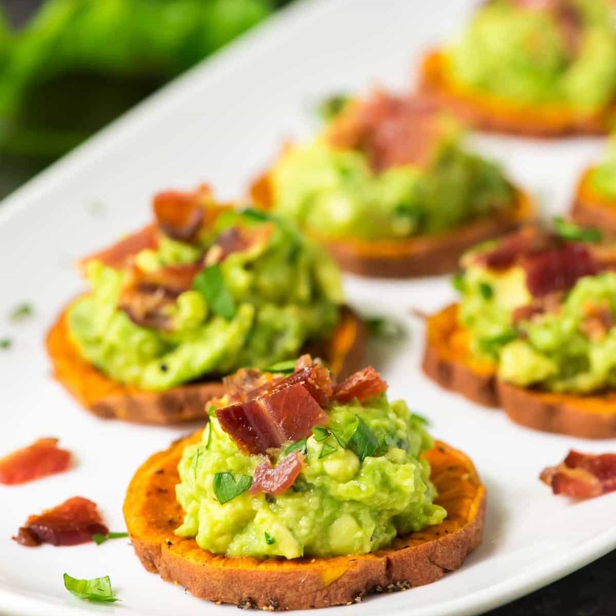 A plate of sweet potato rounds topped with mashed avocado, bacon bits, and fresh parsley.