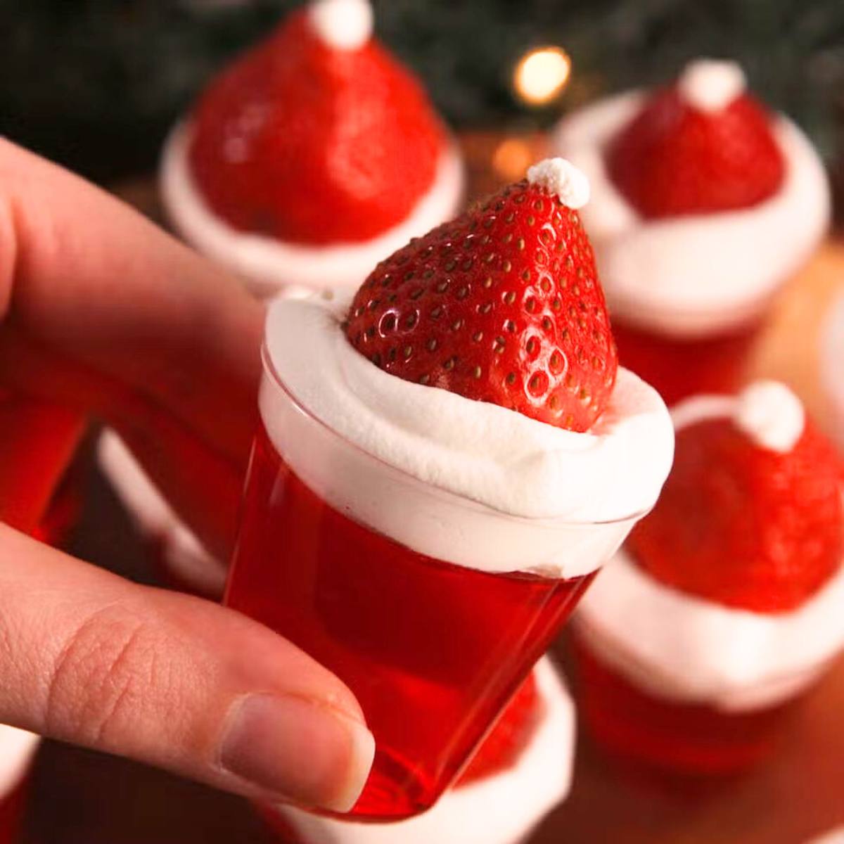 A hand holding a festive Christmas dessert. The dessert is a clear plastic cup filled with red Jell-O, topped with whipped cream and a strawberry. The strawberry is decorated with a small piece of green fruit to resemble a Santa hat.