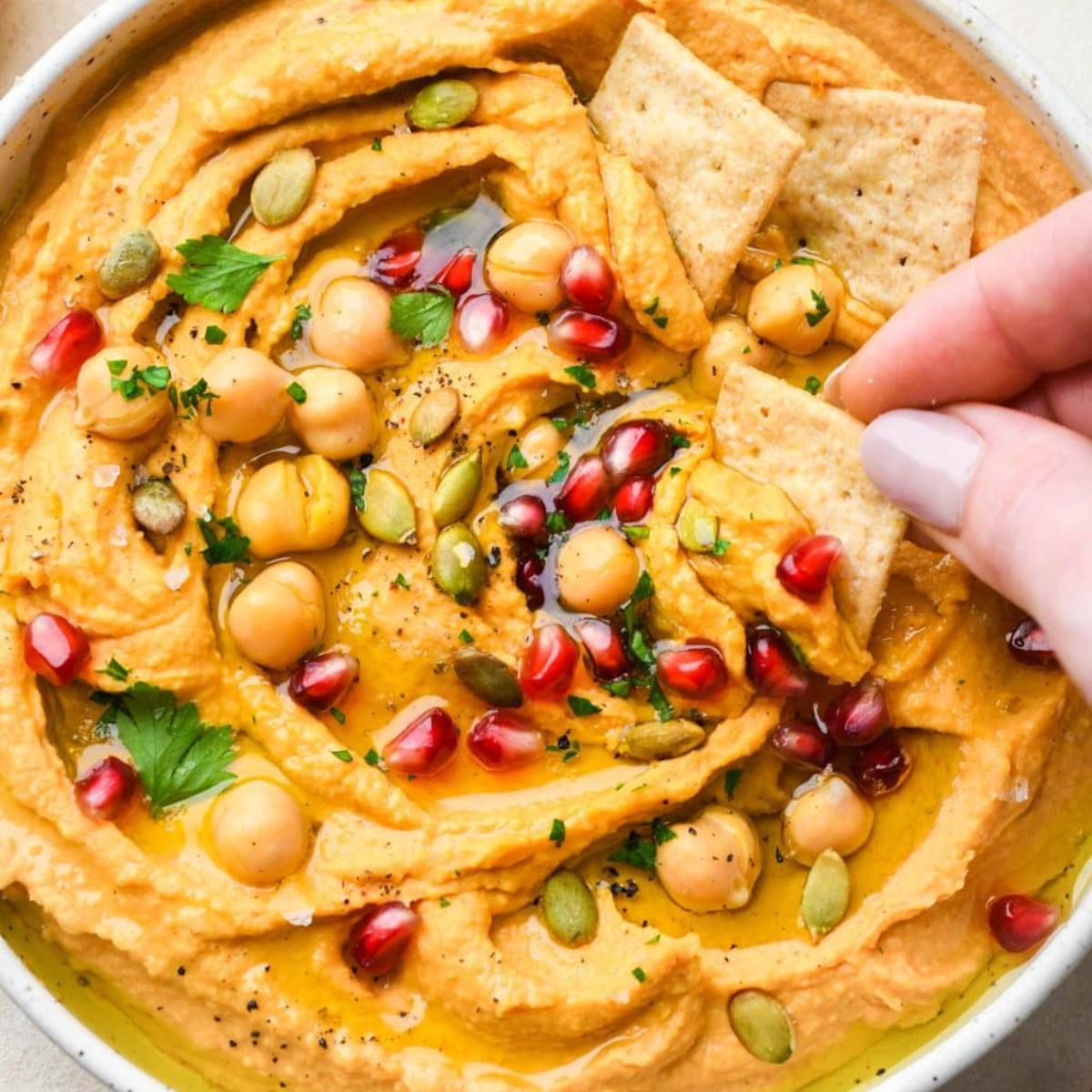 A bowl of creamy sweet potato hummus topped with pomegranate seeds, chickpeas, pumpkin seeds, and fresh parsley. A hand is dipping a cracker into the hummus.