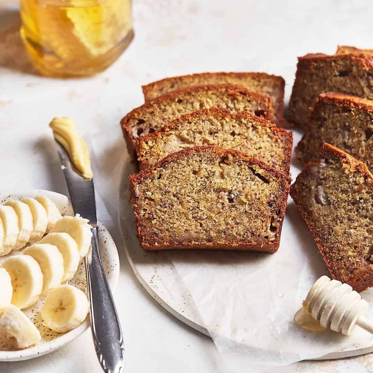 A loaf of freshly air fried banana bread sliced and arranged on a plate. There are sliced bananas, a pat of butter, a honey dipper, and a honey jar on the side. The bread looks moist and delicious.