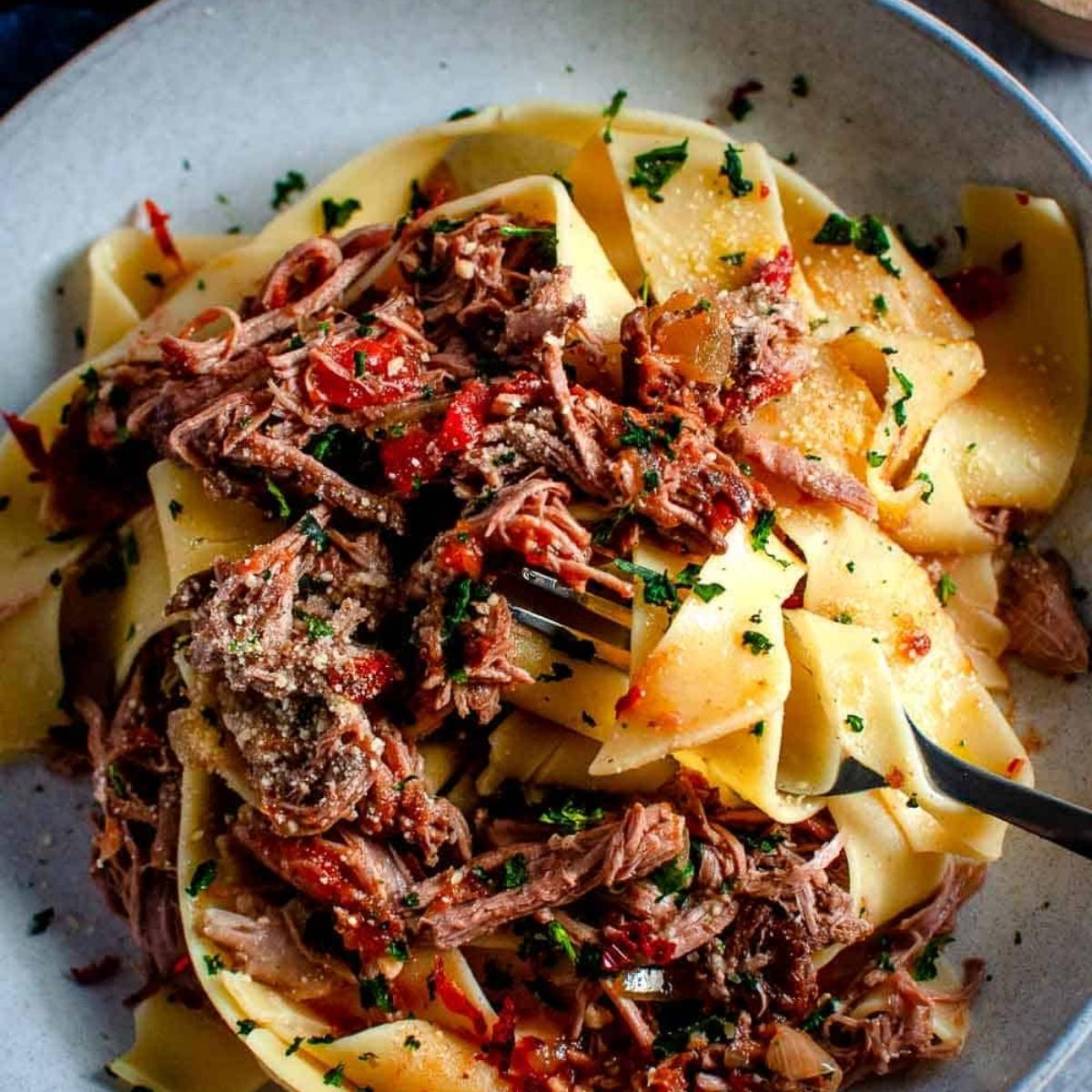 A bowl of pappardelle pasta tossed with a rich, flavorful ragu sauce made with tender shredded meat. The dish is garnished with fresh parsley.