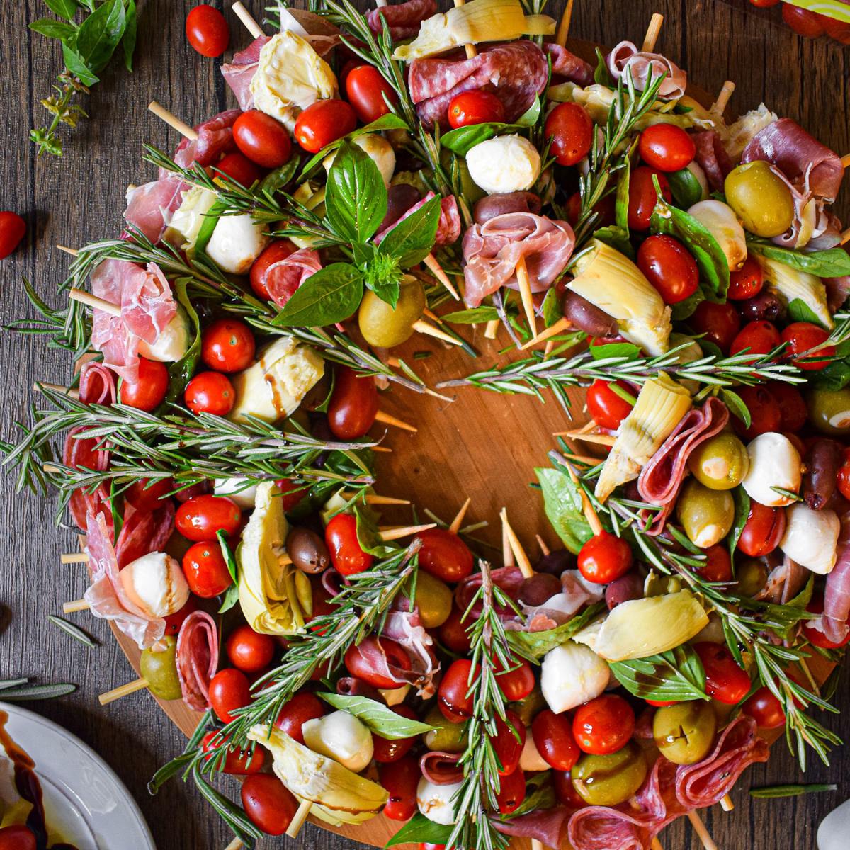 A festive antipasto wreath featuring skewers with cherry tomatoes, mozzarella, salami, olives, and artichoke hearts. The wreath is adorned with fresh rosemary and basil.