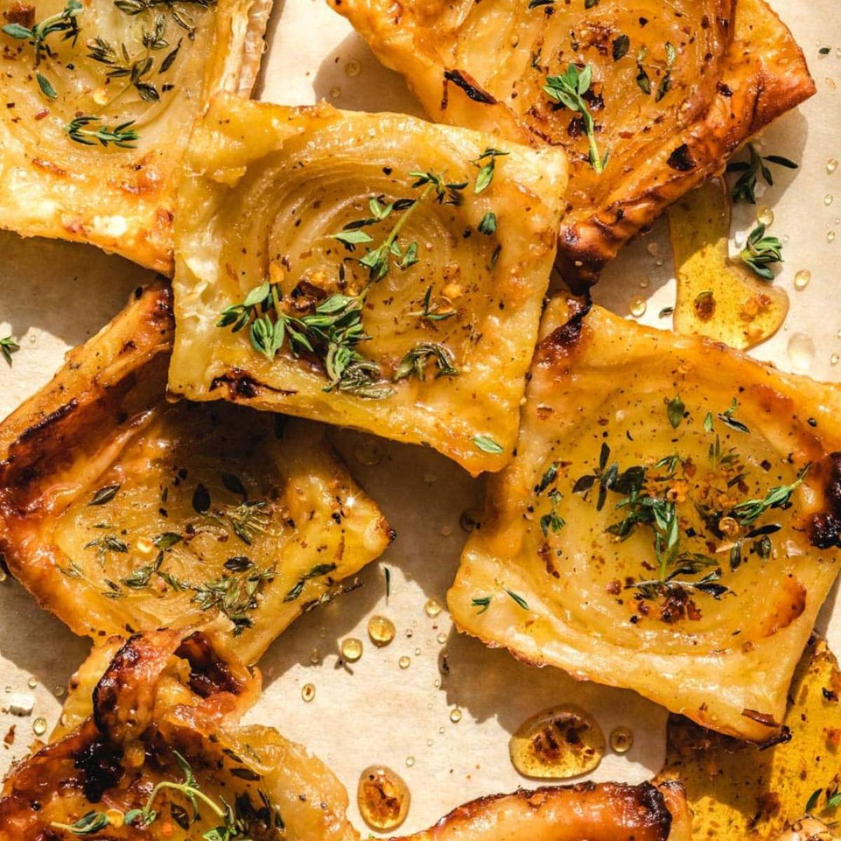 A close-up of several golden-brown puff pastry tarts filled with caramelized onions and topped with fresh thyme sprigs. The tarts are arranged on a parchment-lined baking sheet, and there are drops of honey visible around them.