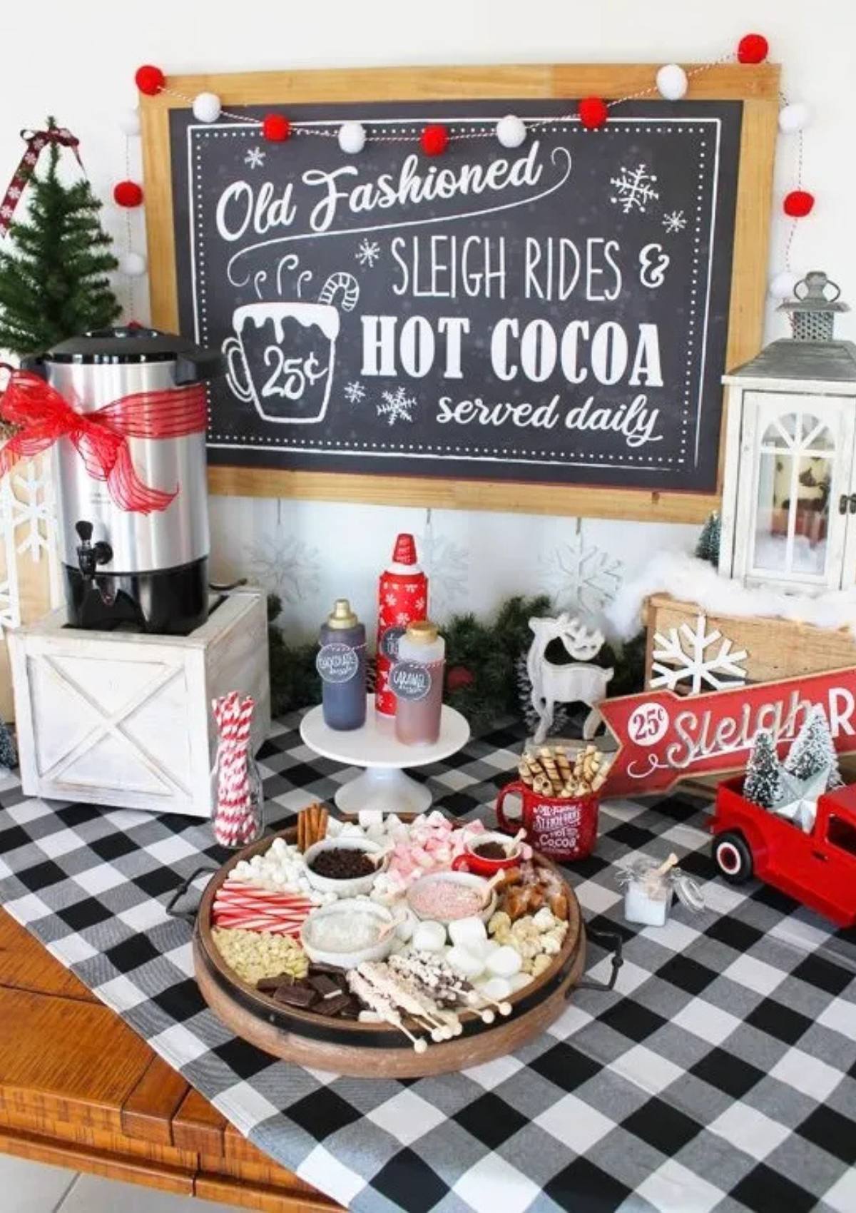 A festive hot cocoa bar with a chalkboard sign that says "Old Fashioned Sleigh Rides & Hot Cocoa Served Daily." The bar includes a variety of toppings, including marshmallows, chocolate chips, candy canes, and sprinkles. There are also mugs, a hot chocolate dispenser, and a red toy sleigh.