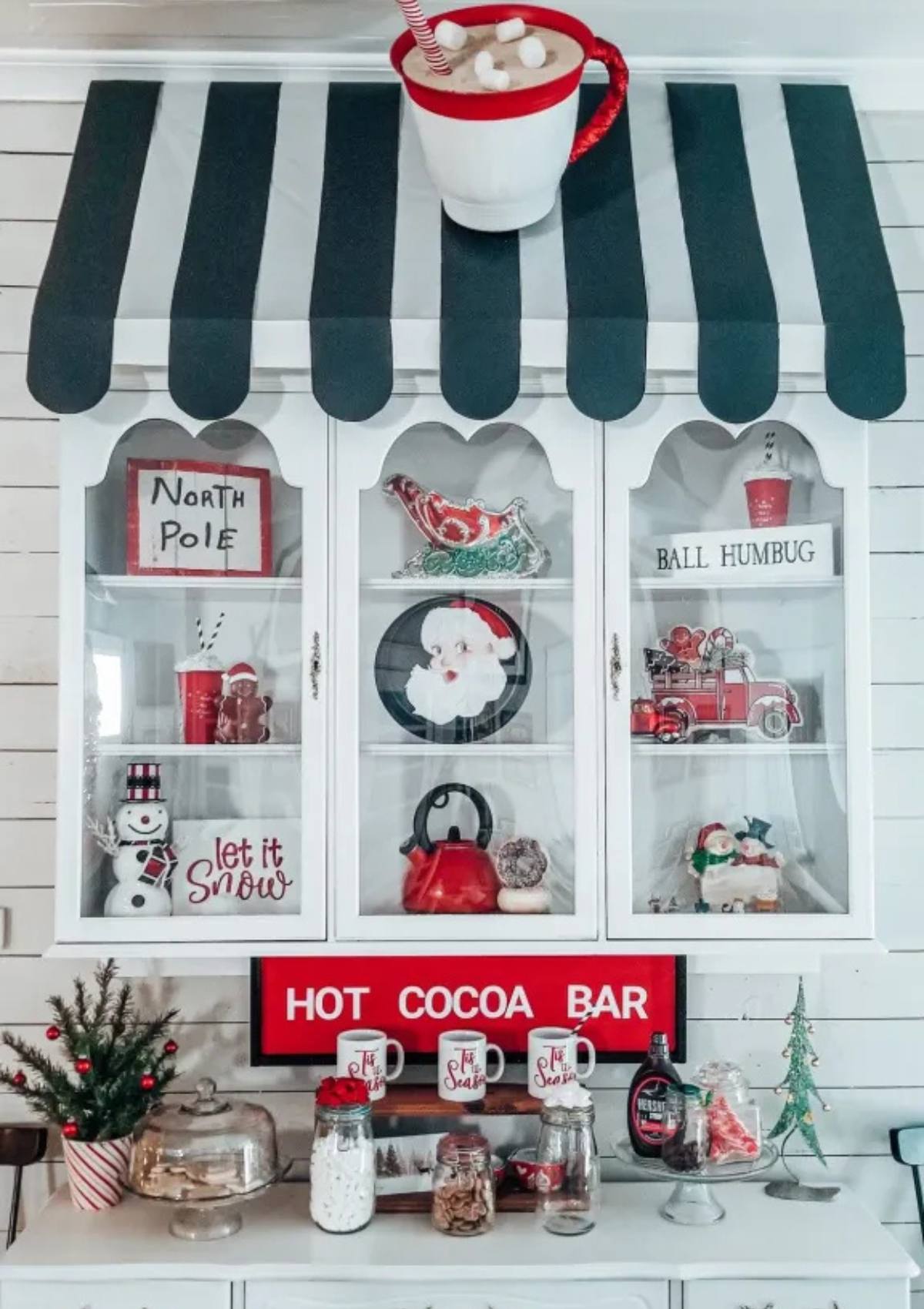 A festive hot cocoa bar setup with a large oversized mug hanging above the counter. The bar is decorated with various Christmas decorations and has a sign that says "Hot Cocoa Bar".